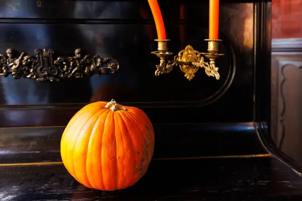 stock image Big ripe orange pumpkin sits atop a vintage black piano with ornate details, accompanied by two lit orange candles in antique holders. The setting evokes a cozy autumn atmosphere Halloween scene.