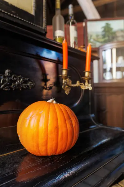 stock image Big ripe orange pumpkin sits atop a vintage black piano with ornate details, accompanied by two lit orange candles in antique holders. The setting evokes a cozy autumn atmosphere Halloween scene.