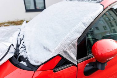 Close-up detail red car with snow-covered windshield protected by reflective cover mat, parked outside residential building during winter. Windscreen window ice frost protection. clipart