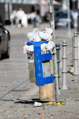 Kalabalık bir çöp tenekesi işlek şehir caddesinde plastik poşetlerle dolu. Sahipsiz atıklar ve çöpler şehir ihmalini ön plana çıkarıyor, sağlık riskleri yaratıyor, yerel çevreyi tehlikeye atıyor ve uygun şekilde imha edilmesi yönünde çağrıda bulunuyor..