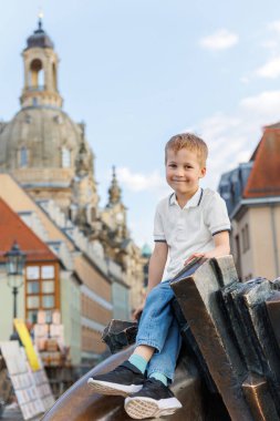 Gülümseyen küçük çocuk, Dresden Almanya 'sında arkasında zarif bir şekilde yükselen Frauenkirche Grand Baroque kubbesine karşı bronz heykelin üzerinde oturuyor. Aile Avrupa şehir seyahati.