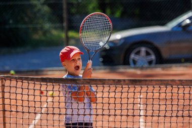 Sevimli küçük mutlu çocuk derste tenis kırmızı kil kortunda topa tek tek vurmaktan zevk alıyor. Yaz sporları kampı. Spor gençlik okulu. Sağlıklı çocuklar aktif yaşam tarzı.