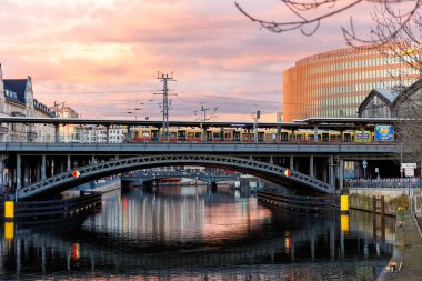 Berlin, Almanya - 25 Şubat 2024: Berlin İstasyonu Friedrichstrasse DB treni dramatik sıcak günbatımı gökyüzüne karşı metal köprüden geçiyor. Akşam Berlin Mitte Spree sahne şehir simgesi.