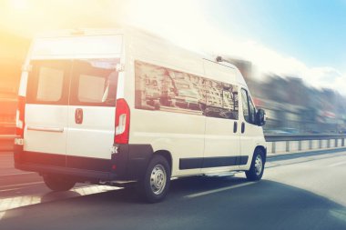 Tanınmayan küçük yolcu minibüsü arka planda şehir manzarası ve gün batımı gökyüzü ile şehir sokak trafiğinde karayolu üzerinde acele. Charter veya servis otobüsü minibüsü hareket bulanıklığı etkisi ile yolda acele.