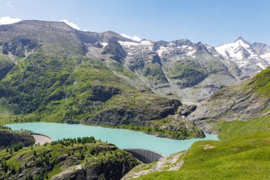 Turkuaz Grossglockner buzul rezervuarı Speicher Margaritzen alp yamaçları ve vadi kenarında beton baraj yapısı olan orman tepeleri. Canlı doğal dağ manzaralı yüksek irtifa gölü.