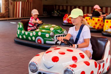 İki çocuk renkli çarpışan arabalara biniyor, gülüyorlar ve birbirlerine bakıyorlar. Geniş pencerelerden gelen noktalı ve güneş ışığına sahip parlak boyalı araçlar neşeli ve eğlenceli bir ortam yaratıyor.