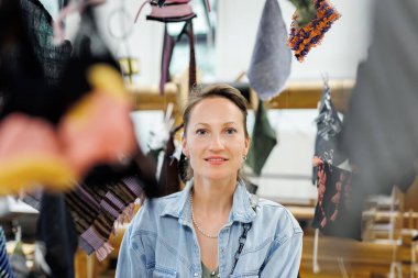 Kumaş örnekleriyle çevrili bir kadın tekstil atölyesinde duruyor. Kot gömlek ve inci kolye giyerken nazikçe gülümsüyor. Renkli dokuma kumaş ipliklerle çerçevelenmiş..