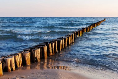 Ahşap dalgalar, yumuşak akşam ışığı altında mavi denize doğru uzanıyor. Yumuşak dalgalar kumlu sahile dokunurken, güneş sıcacık ahşap direklere ve parıldayan suya yansıyor..