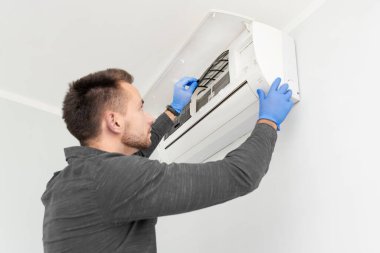 Bir adam klimayı tamir ediyor. O eldiven ve mavi bir gömlek giyiyor.