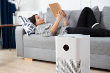 Kadın kanepede kitap okuyor. Kanepenin yanında beyaz bir hava temizleyici var.