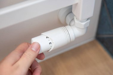 Kişi radyatörün sıcaklığını ayarlıyor. Radyatör beyaz ve üzerinde düğmesi var.