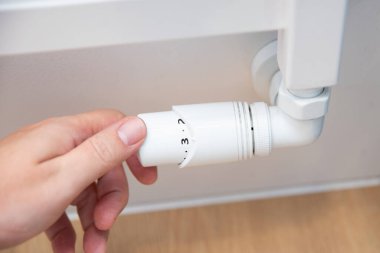 Kişi radyatörün sıcaklığını ayarlıyor. Radyatör beyaz ve üzerinde düğmesi var.