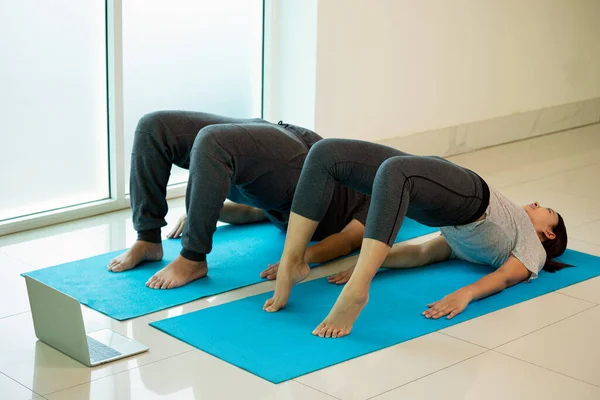 Two Asian young male and female adult lying for practice Yoga pose Bridge or Setu Bandha Sarvangasana in gym near window