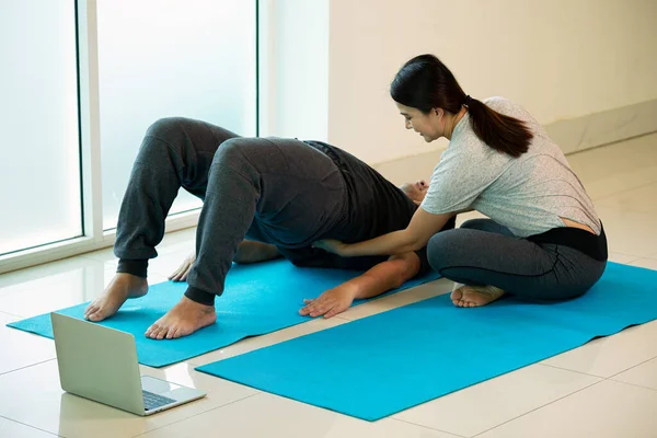 Two Asian young male lying and female support back for practice Yoga pose Bridge or Setu Bandha Sarvangasana in gym near window