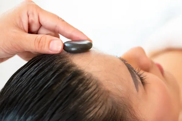 a close up therapist hand palcing black spa stone on customer forehead laying down on bed; Holistic Health and Well-Being Healing Hand with Hot Stone