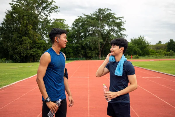 Two Asia male Athlete Drinking Water talking wiping face on Track together; Mineral Water Stay Active and Hydrated Break