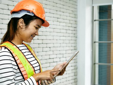 Turuncu şapkalı bir kadın müteahhit, beyaz tuğladan duvarı olan bir ofisteki tabletteki dijital planları dikkatle incelerken gülümsüyor.