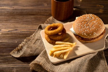 Susamlı çörek çizburger, soğan halkaları ve tahta tahtada servis edilen patates kızartması.