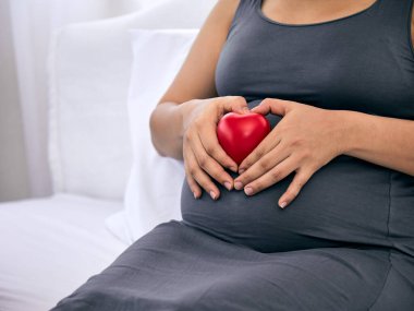 Huzurlu bir ortamda hamile bir kadın, karnında şefkatle kırmızı bir kalp taşıyor, sevgiyi ve doğmamış çocuğuyla olan bağını simgeliyor.