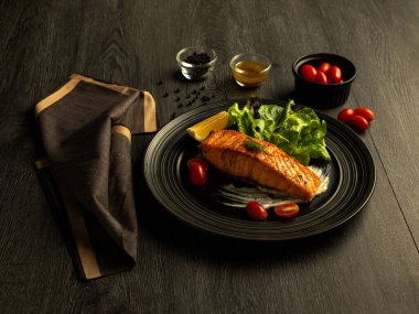 Lezzetli ızgara somon filetosu taze salata ve vişneli domates ile servis edilir, ahşap arka planda şık koyu tabakta sunulur.