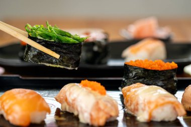 Bir parça suşiyi çeşitli suşi tabağının üzerinde yosun salatasıyla birlikte tutan yemek çubuklarının yakın çekimi, çeşitli nigiri ve roe çeşitleri içerir.