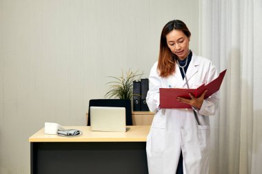 Beyaz önlüklü Asyalı kadın doktor tıbbi ofiste duruyor, hasta kayıtlarını panoda inceliyor, tıbbi profesyonelliği ve hasta bakımını vurguluyor.