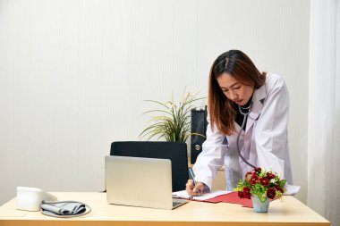 Steteskoplu Asyalı kadın doktor, tıp bürosundaki masasında dizüstü bilgisayar ve tıbbi malzemelerle notlar yazıyor.