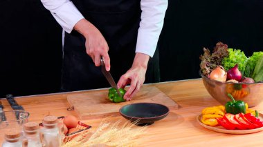 Siyah önlüklü şef, taze sebze ve malzemelerle çevrili ahşap kesme tahtasına yeşil bir dolma biber dilimliyor.