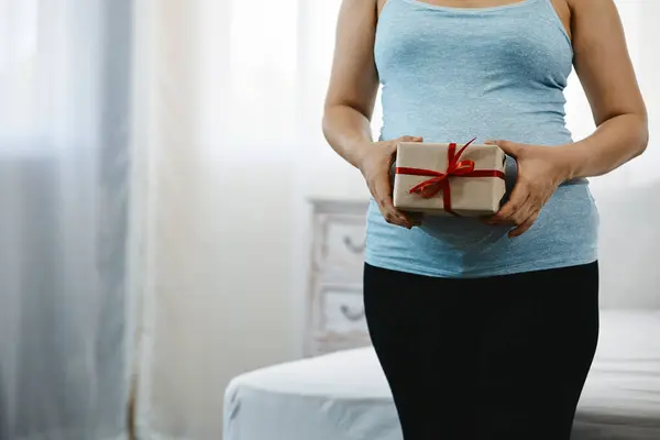 stock image Pregnant woman in blue tank top holding gift box with red ribbon, standing indoors near bed, softly lit room with copy space