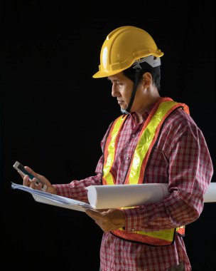 Sarı şapka ve güvenlik yeleği giyen erkek inşaat işçisi. Elinde karanlık arka plana karşı planlar ve akıllı telefon var.