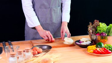 Gri önlüklü genç şef, mutfak tahtasının üzerinde yumurta ve sebzeler de dahil olmak üzere malzemeler hazırlıyor.