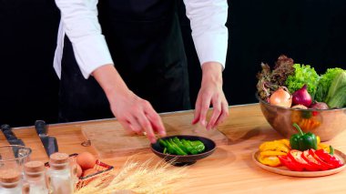 Siyah önlüklü genç şef taze sebze ve malzemelerle çevrili yeşil dolma biberi kasede dikkatlice dilimledi.