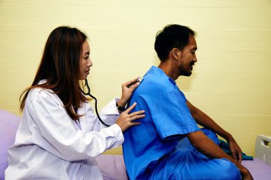 Kadın doktor hastane yatağında oturan hastayı muayene etmek için steteskop kullanıyor. Rutin kontroller sırasında hastanın sırtını kontrol ediyor.