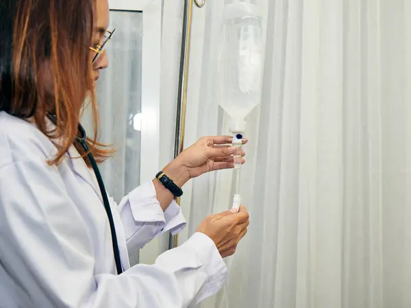 Beyaz önlüklü kadın doktor, serum serumunu hastane odasında ayarlıyor ve uygun sıvı yönetimini sağlıyor.