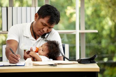 Çoklu görev yapan baba bebeğini besliyor, telefonla konuşuyor ve masa başında yazıyor, aynı anda hem işi hem de ebeveynliği yönetiyor.