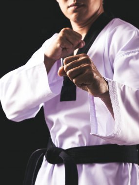 One martial artist wearing white uniform and black belt assumes fighting stance on black background clipart