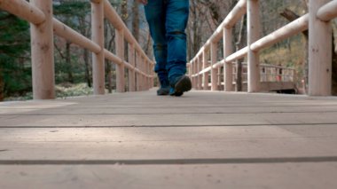 Ahşap köprüde yürürken, botlarına ve hareketlerine odaklanmış, yakın açılı bir çekim.