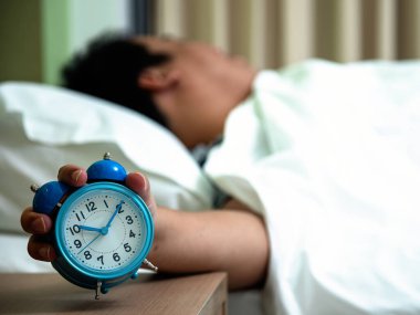 Yatakta yatan bir adam, elinde mavi alarmlı saati, sabah rutinlerini ve uyanma alışkanlıklarını simgeliyor.