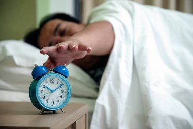 Yatakta uzanmış bir adam yatağın başucundaki mavi saati uyutmak için elini uzatıyor, tipik bir sabah rutini.