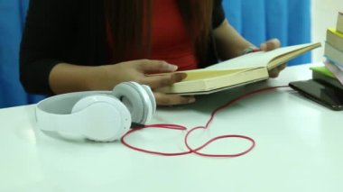 Beyaz masanın üzerinde elinde kitap tutan ve okuyan bir kadın, beyaz kulaklıklar ve kırmızı kablo yanında kitap ve akıllı telefon yığını