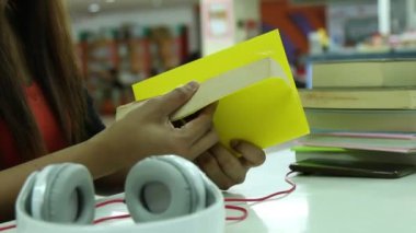 El ele tutuşmalar ve açık sayfalı sarı kitap çevirmeler, beyaz kulaklıklar, ve kütüphanedeki bir yığın kitap.