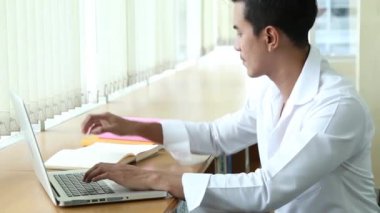 Beyaz günlük gömlek giyen genç Asyalı erkek araştırmacı kütüphanede dizüstü bilgisayarla çalışıyor, yanında kitaplar ve notlar var.