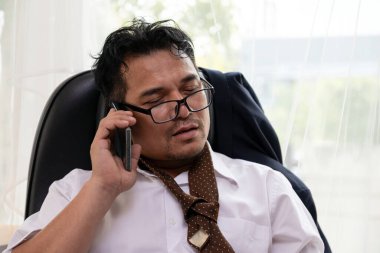 Gevşek kravatlı ve eğik gözlüklü bitkin işadamı telefonda konuşurken sandalyeye yaslanıyor, stresli ve fazla çalışmış görünüyor.