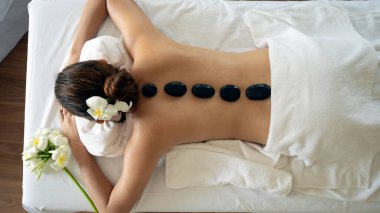 Genç bir kadın, beyaz çiçeklerle süslenmiş masaj masasında yatarken sırtında siyah bazalt taşlarıyla sıcak taş masajından hoşlanır.