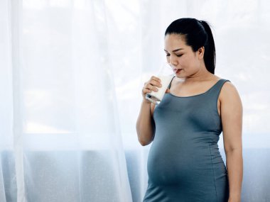 Mavi elbiseli Asyalı hamile kadın pencerenin yanında duruyor, hamileliği boyunca bir bardak süt içiyor.
