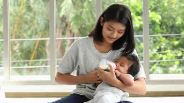 Genç anne evde otururken bebeğini sevgiyle süt şişesiyle besliyor, sakin ve besleyici bir an yaratıyor.