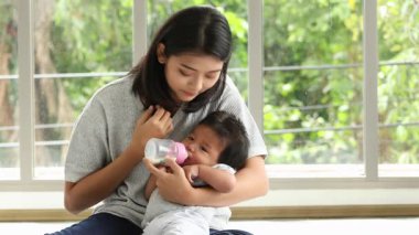 Genç bir anne, bebeğini nazikçe bir şişeyle beslerken parlak bir pencerenin yanında oturuyor ve ona bakıcılık yapıyor.