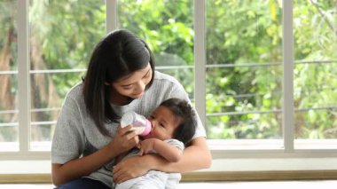 Bir anne, güneşli bir pencerenin yanında otururken bebeğini sevgiyle besler ve huzurlu bir aile anı yaratır.