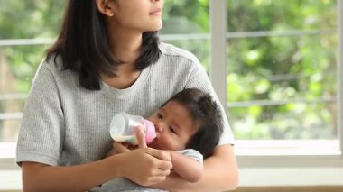 Sevgi dolu bir anne bebeğini süt şişesiyle tutup besler. Pencere kenarında huzurlu ve besleyici bir an yaratır.