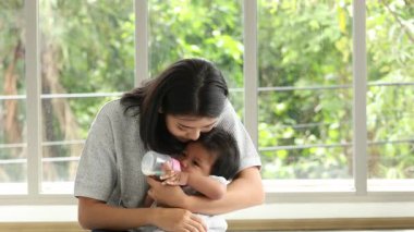 Sevgi dolu bir anne bebeğini süt şişesiyle tutup besler. Pencere kenarında huzurlu ve besleyici bir an yaratır.